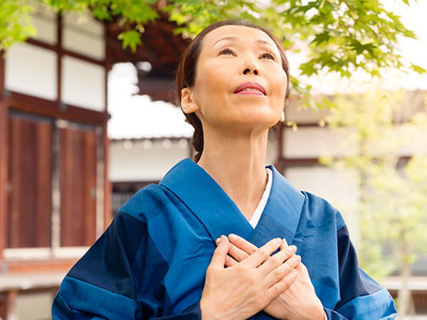 A mature Japanese woman wearing a blue kimono has her hands resting over her heart in a healing gesture as she looks up toward the sky.